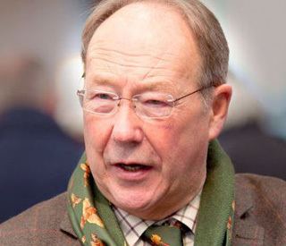 David Barby talking to someone while wearing a brown coat, white and brown long sleeves, green scarf, green necktie, and eyeglasses
