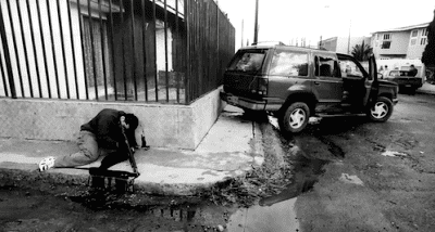 David Barron Corona holding a gun and leaning on the fence with blood on the floor near the street corner while wearing a jacket and pants