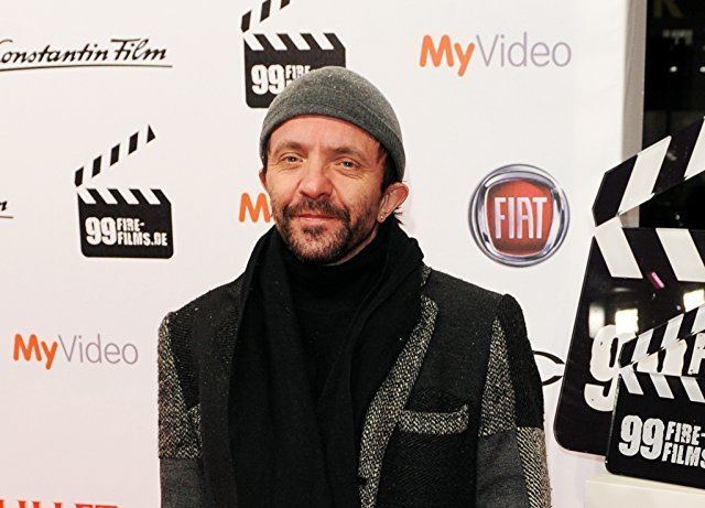 David Bennent with a tight-lipped smile, mustache, and beard while wearing a black and gray coat, black scarf, and a gray beanie