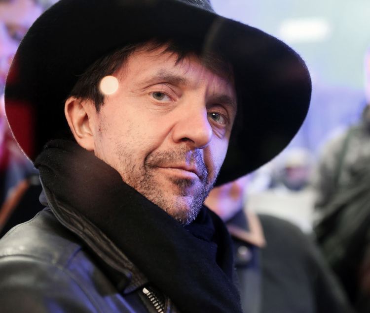 David Bennent with a tight-lipped smile, mustache, and beard while wearing a black jacket, black scarf, and a black hat