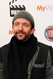 David Bennent with a tight-lipped smile, mustache, and beard while wearing a black and gray coat, black scarf, and a gray beanie