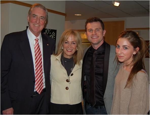 David Campbell wearing a suit and a black tie with a man wearing a suit and a red striped tie, a woman with blonde hair, wearing a yellow shirt and a necklace, and a girl wearing a brown sweatshirt.