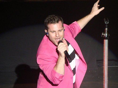 David Campbell wearing an earpiece, a pink coat, and a white and black shirt while holding a microphone.