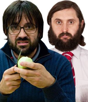 David Earl peeling a green vegetable using a butter knife with a disgusted face, mustache, and beard while behind him is Joe Wilkinson with a serious face, mustache, and beard. David is wearing a light-blue shirt under and a blue jacket while Joe is wearing a white long sleeve and a red and white striped necktie
