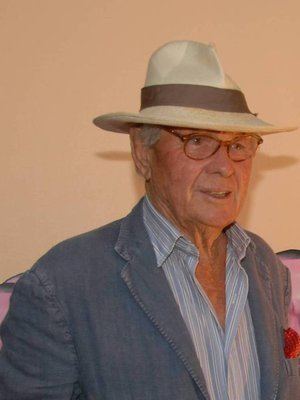David Hamilton (photographer) wearing a white striped shirt, a gray suit, a pair of eyeglasses, and a brown hat