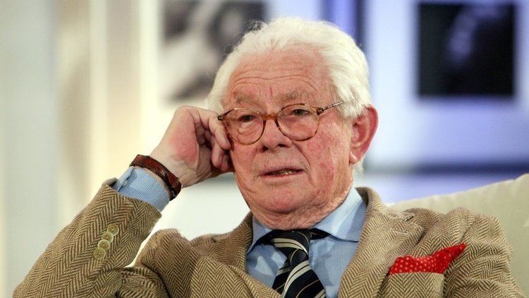 David Hamilton (photographer) wearing a blue long-sleeved shirt, a brown suit, and a pair of eyeglasses while sitting