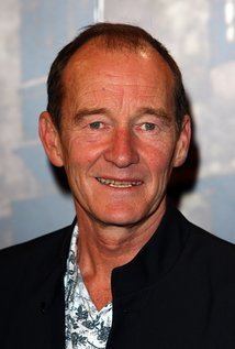 David Hayman smiling during an event and wearing a white, floral designed shirt underneath a black suit.