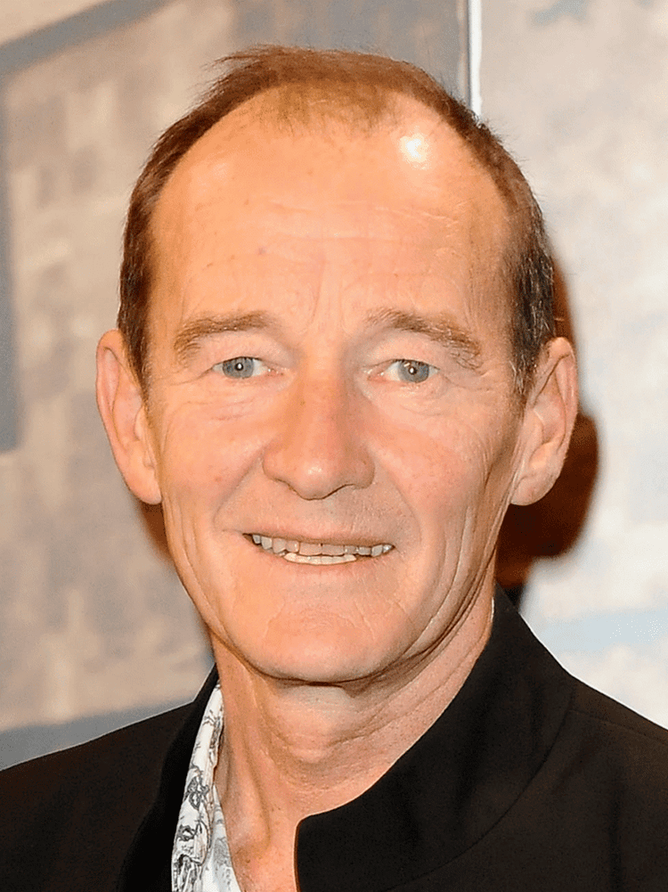 David Hayman smiling during an event and wearing a white, floral designed shirt underneath a black suit.