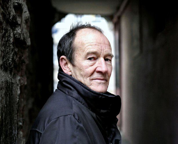 David Hayman posing and wearing a black collared coat.