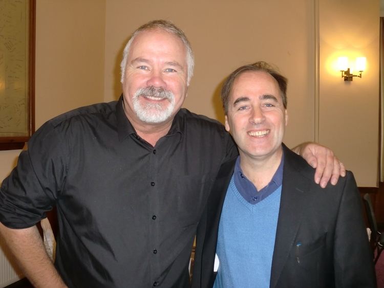 David Lonsdale smiling with gray hair, beard, and mustache and embracing the shoulder of the man beside him, wearing a black shirt