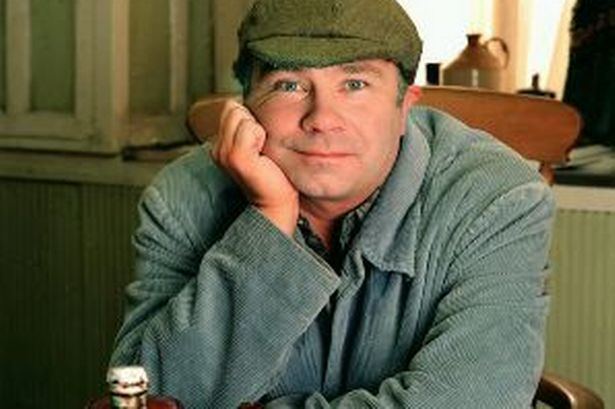 David Lonsdale smiling, supporting his chin by his hand and wearing a blue knitted jacket and green cap