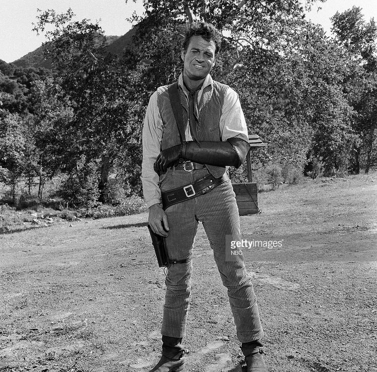 In black and white, in a field with a small well and trees at the back, David McLean is smiling, standing, legs apart, left arm in a splint with sling to his right shoulder, has black hair, wearing a white long sleeve, a black gray corduroy vest, black belt, a belt with gun holster, gray corduroy pants, and gray shoes.