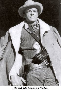 In black and white, David McLean is serious, standing with his right hand on the gun holster, left arm in a splint with a sling to his right shoulder, wearing a white cowboy hat, white polo under corduroy gray vest, corduroy gray pants, a belt with gun holster, and a gray long coat.
