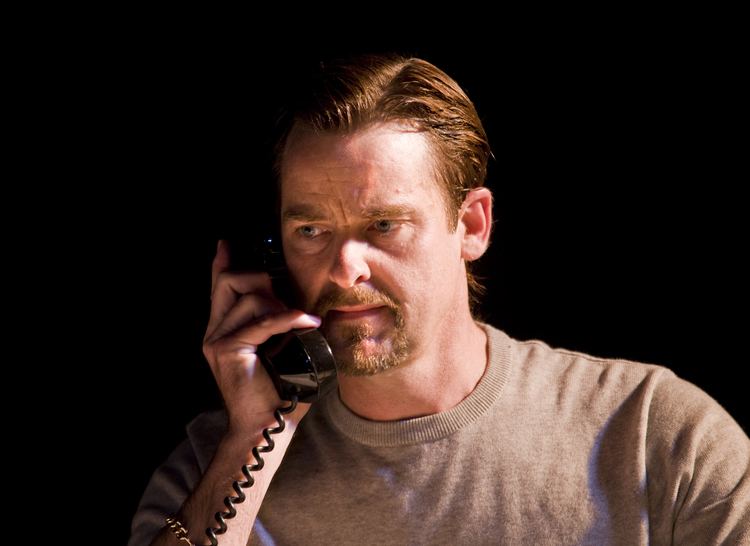David Michaels answering a phone and looking serious with a mustache and beard while wearing a gray shirt
