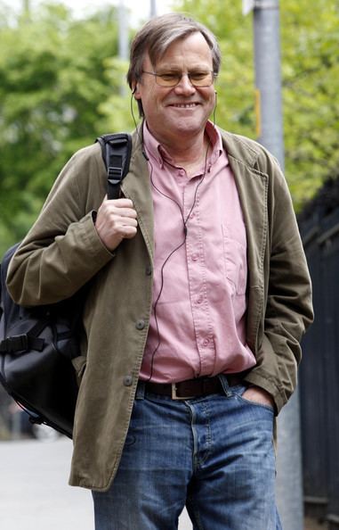 David Neilson smiling while carrying a black bag and wearing a brown coat, pink long sleeves, denim pants, eyeglasses, and an earphone