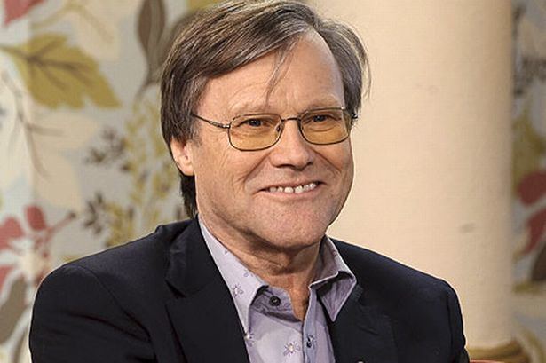 David Neilson smiling while wearing a black coat, blue long sleeves, and eyeglasses