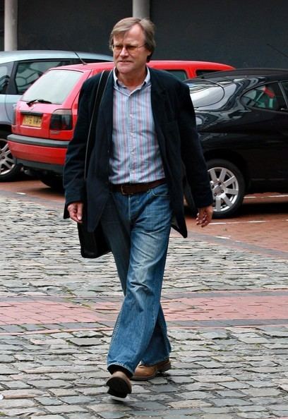 David Neilson looking at something while walking and wearing a black coat, blue striped long sleeves, denim pants, and brown shoes