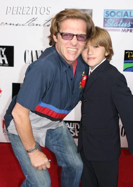 David Rainey smiling with his son, Bogart Rainey while he is wearing a blue and red polo, red inner shirt, denim pants, and sunglasses