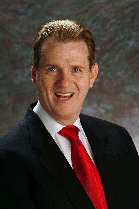 David Ring is happy, mouth half open, in front of a backdrop, has brown hair, wearing a white polo with red necktie under a black coat.