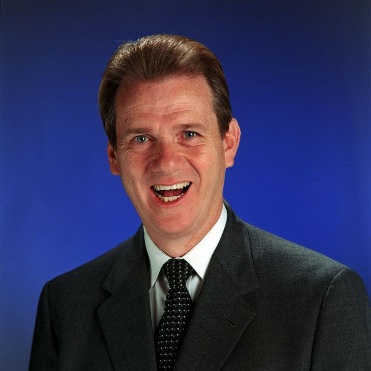 David Ring is happy, mouth half open, in front of a Blue background, has brown hair, wearing a white polo with black dotted necktie under a black coat.