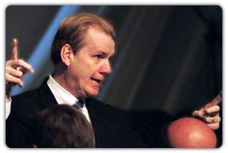 David Ring is serious, mouth half open, in front of people in a room, hands pointing up, speaking, has brown hair, wearing a white polo with black dotted necktie under a black coat.
