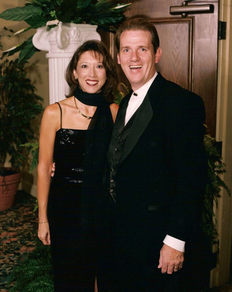 In a room with yellow walls and a large pillar centerpiece with large leaves and plants on top. From left, Karen Ring is smiling, standing and has long black hair wearing a black scarf, earrings bracelet, gold ring, gold necklace, and a noodle strap black dress, at the right, David ring is happy, standing, mouth half open, right hands over Karen’s waist, has brown hair wearing a gold ring, white long sleeve polo under a black coat.