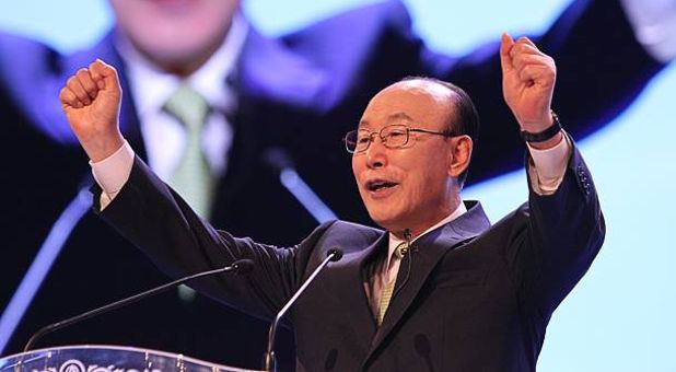 David Yonggi Cho speaking to an audience on stage and wearing a dark brown coat and eyeglasses.