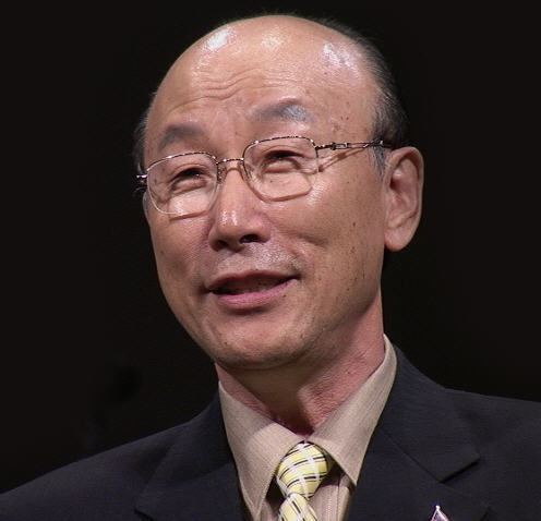 David Yonggi Cho smiling and wearing a black coat over a light brown suit and yellow tie and eyeglasses.