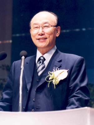 David Yonggi Cho smiling on stage and wearing a blue formal attire over suit with a striped tie and a yellow corsage.