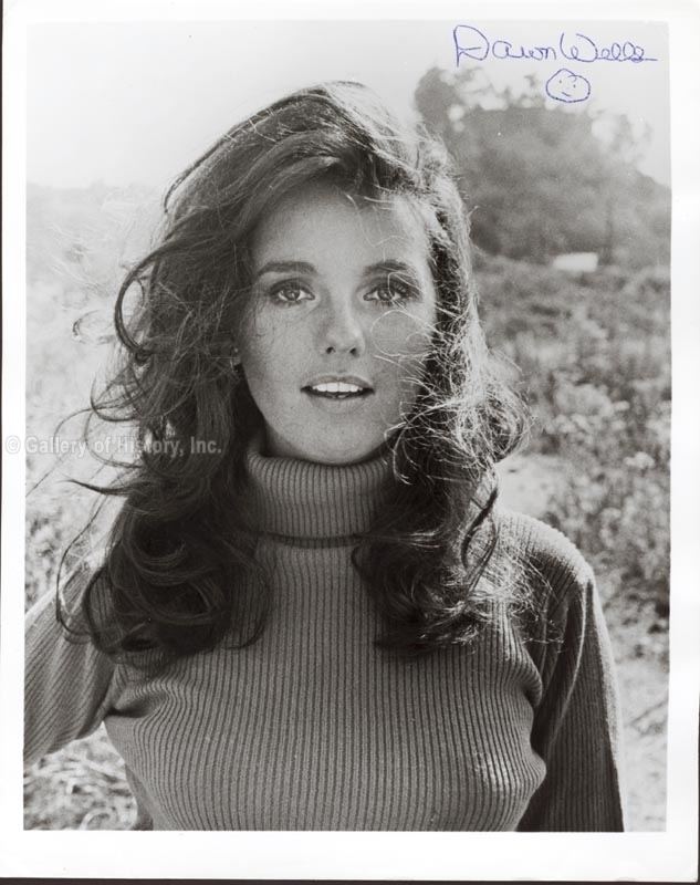 Dawn Wells smiling with her wavy black hair in an old photograph with “Dawn Wells” written on it. She is wearing an earring and a high neck long sleeve