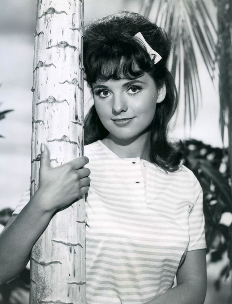 Dawn Wells smiling while hugging a tree with her right hand in an old photograph, she has black short hair with bangs and a white ribbon clip and wearing a striped shirt
