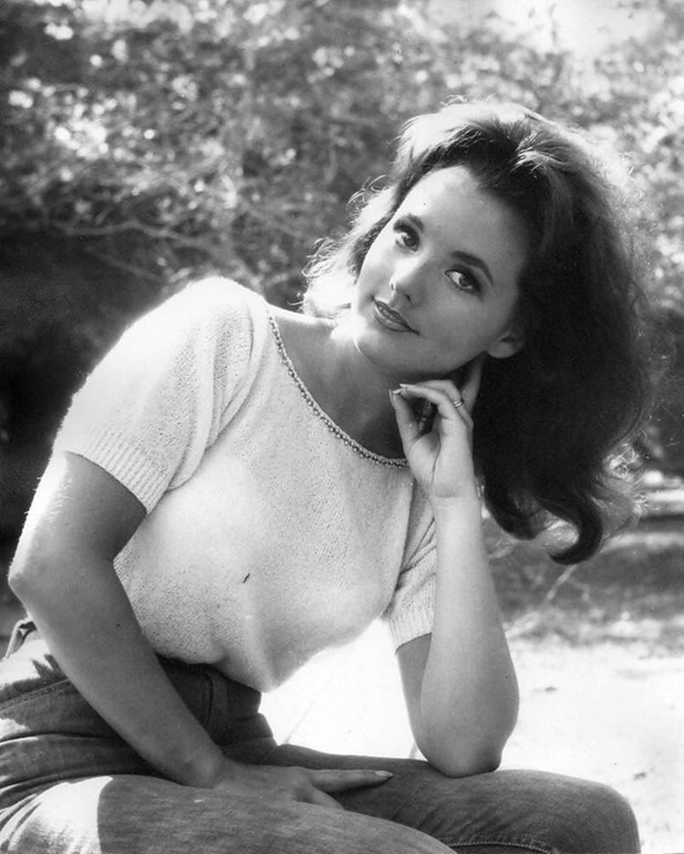 Dawn Wells smiling and sitting while leaning on her two left fingers with trees on her background in an old photograph, she has black hair wearing a ring on her left finger, white blouse and denim jeans