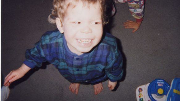 On the left, Jeffrey Baldwin is happy, standing heads up looking to his left with a kid's hand on his back. has brown hair, wearing a blue checkered long sleeve polo. At the bottom right is a white with blue car toy.