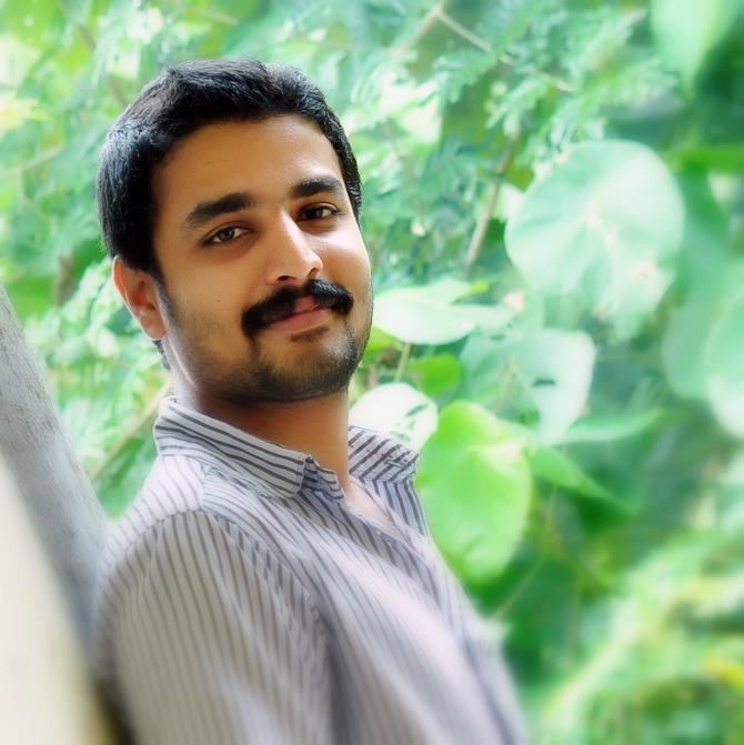 Deepak Parambol smiling, with beard and mustache, and wearing a striped polo shirt while leaning on the wall.