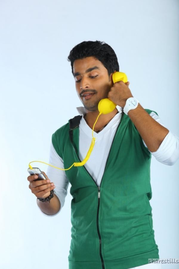 Deepak Parambol with beard and mustache, wearing a white long sleeve shirt and green vest while holding a yellow telephone connected to a mobile phone.