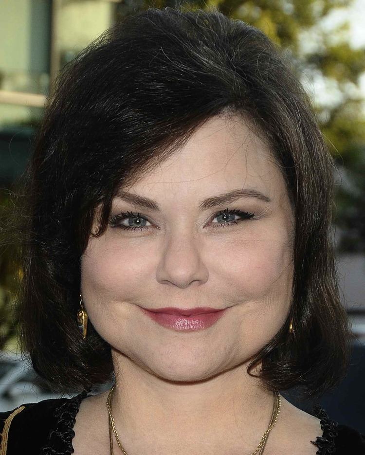 Delta Burke smiling while wearing a black blouse, gold necklace, and gold earrings