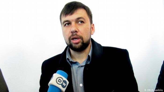 Denis Pushilin is talking in front of a microphone while looking afar, with a beard and mustache, wearing a black coat over gray long sleeves.