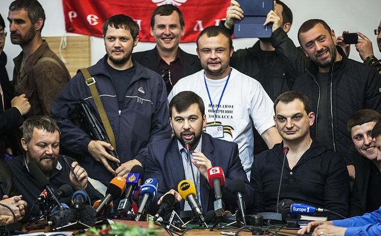 Denis Pushilin (center) talking in front of the media during a conference with his colleagues. Denis with beard and mustache, wearing a dark blue coat over light blue long sleeves.