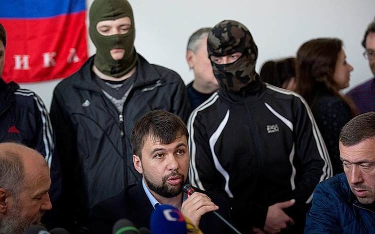 Denis Pushilin (center) talking in front of the media during a conference with his colleagues. Denis with beard and mustache, wearing a dark blue coat over light blue long sleeves while some of his colleagues are wearing masks.