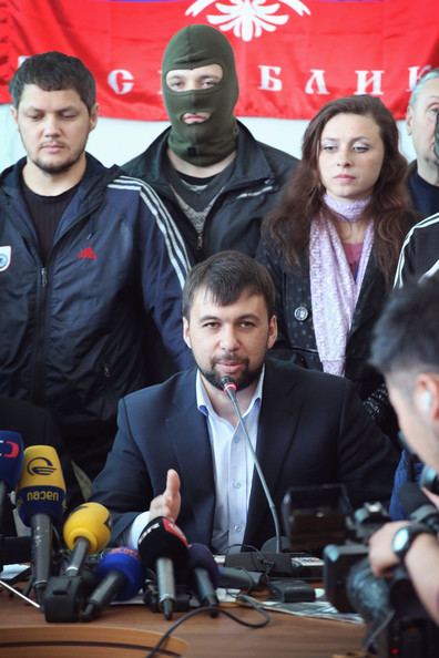 Denis Pushilin (center) talking in front of the media during a conference with his colleagues. Denis with beard and mustache, wearing a dark blue coat over light blue long sleeves while one of his colleagues is wearing a mask.