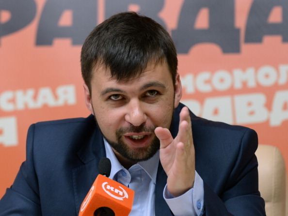 Denis Pushilin is talking in front of a microphone while moving his hand, with beard and mustache, wearing a blue coat over light blue long sleeves.
