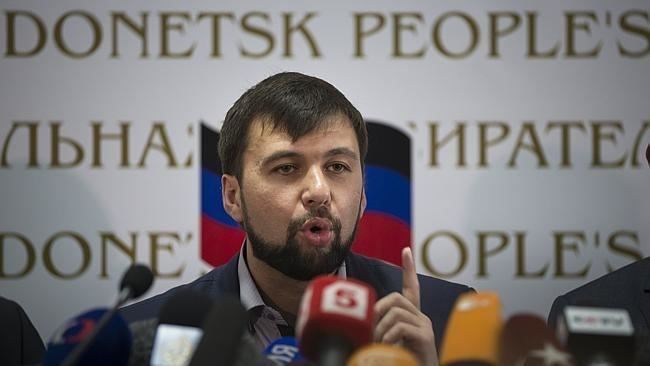 Denis Pushilin is talking in front of the media while pointing his finger with the flag of the Donetsk People's Republic in the background. Denis with beard and mustache, wearing a dark blue coat over gray long sleeves.