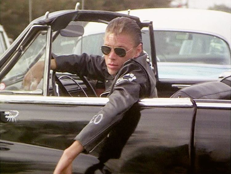 Dennis Cleveland Stewart in "Grease" movie driving a black car, wearing sunglasses and a black jacket.