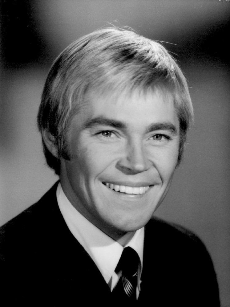 Dennis Cole with a smiling face, wearing a black suit, a tie, and white long sleeves.