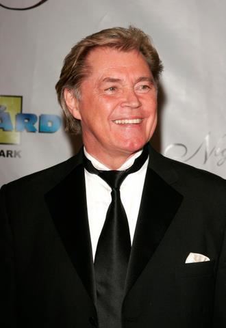 Dennis Cole with a smiling face, wearing a black suit, a black tie, and white long sleeves.