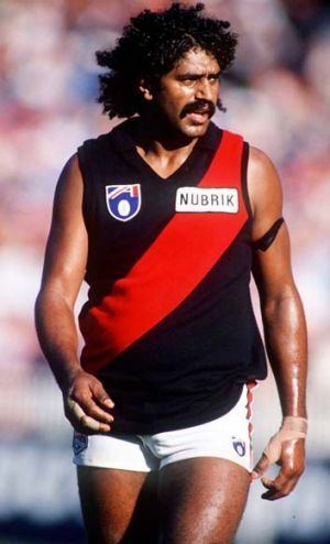 Derek Kickett with curly black hair and a mustache, wearing a red and black jersey and white shorts.