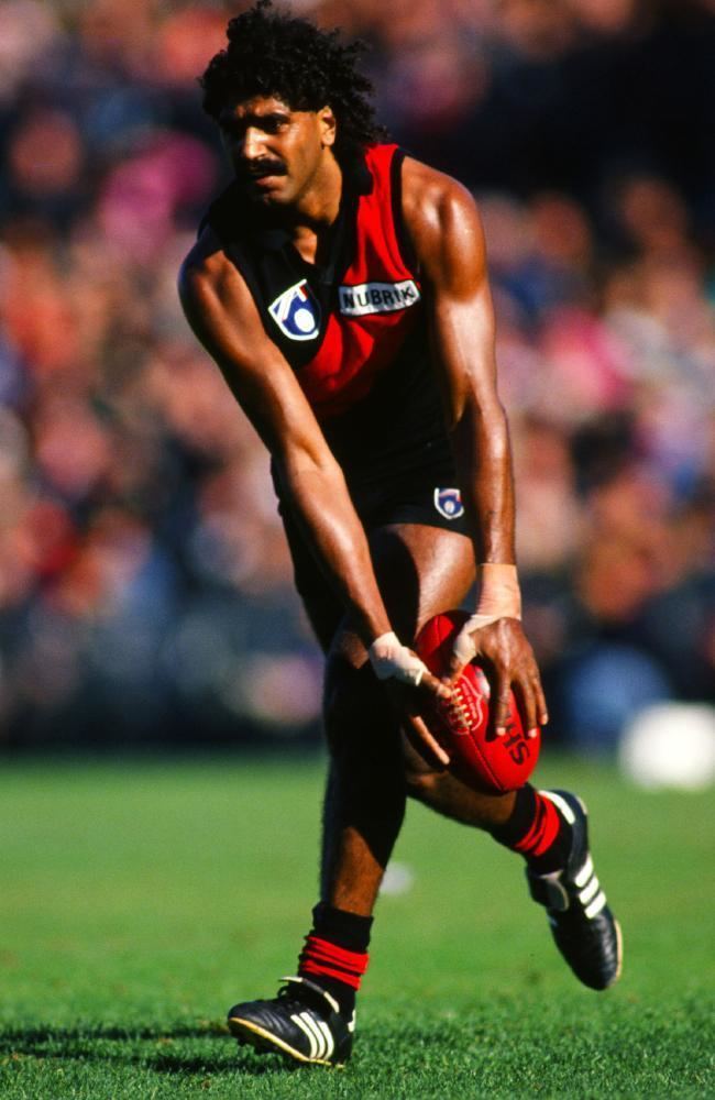 Derek Kickett with curly black hair, wearing a red and black jersey, black shorts, black and white shoes, red and black socks, and holding a football ball.