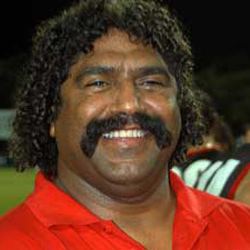 Derek Kickett smiling, with a mustache and curly black hair and wearing a red shirt.