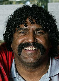 Derek Kickett smiling with a mustache, with curly black hair, wearing a gray polo shirt and red jacket.