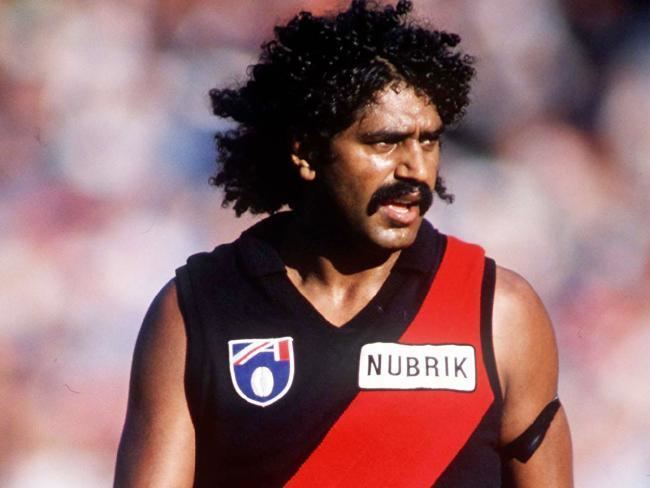 Derek Kickett with curly black hair and a mustache and wearing a red and black jersey.